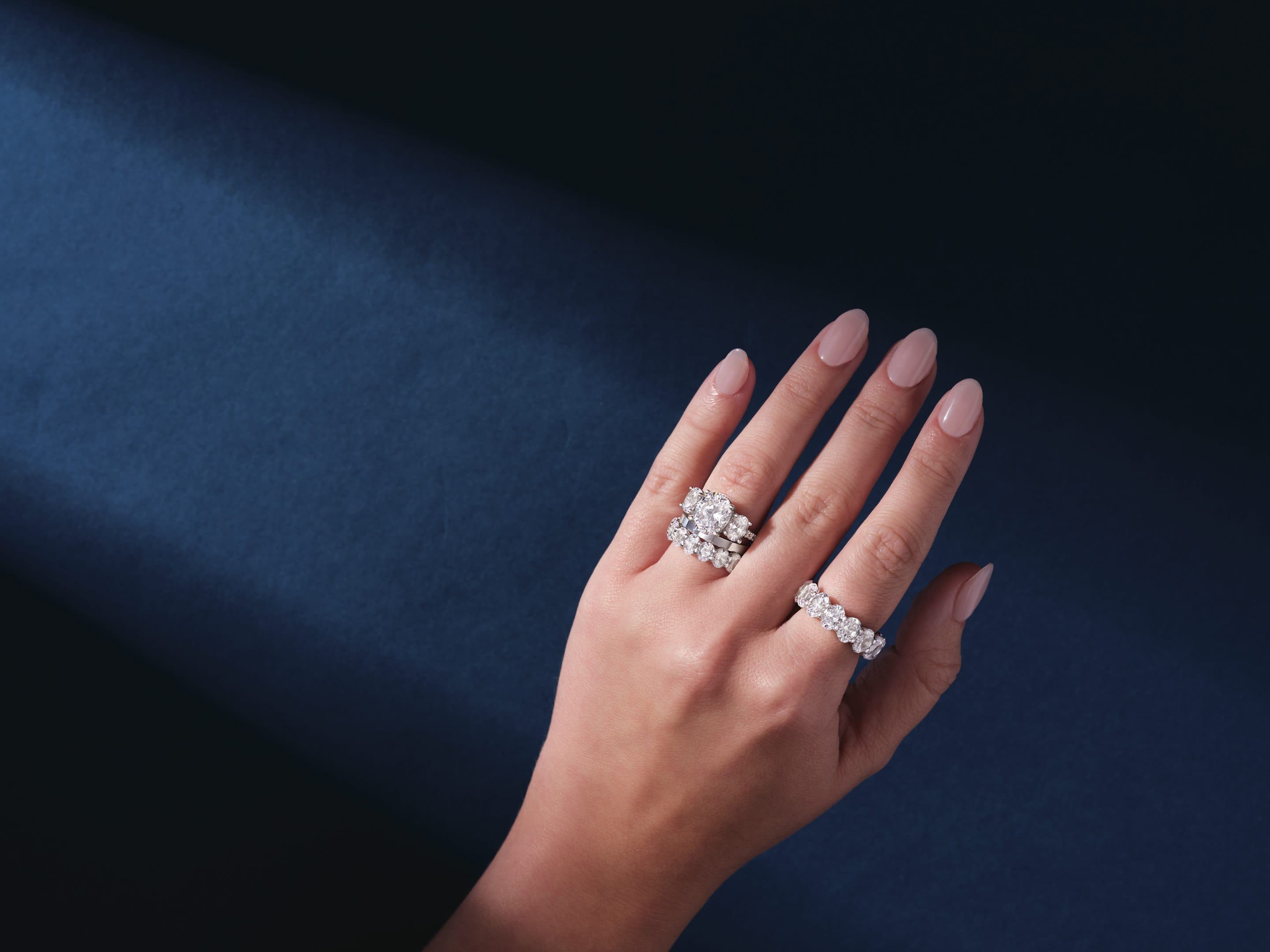 Hand wearing a diamond engagement ring and eternity band against dark navy fabric